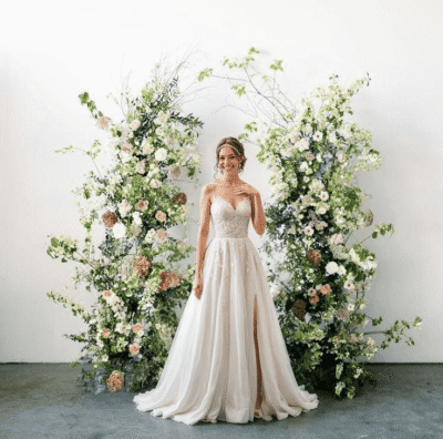 ride in a lace wedding dress standing between tall asymmetrical greenery floral arrangements with white and blush flowers in a minimalist indoor setting.