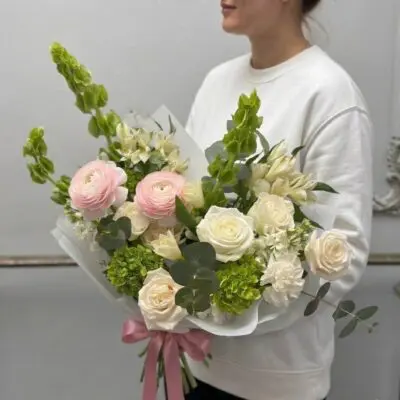 Hand-tied bouquet with blush pink ranunculus, white roses, fresh green blooms, delicate white flowers, eucalyptus, and tall green stems wrapped in white paper with a pink ribbon.