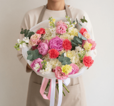 Pastel bouquet with pink peony-style flowers, cream blooms, coral accents, and eucalyptus wrapped in white paper with ribbon