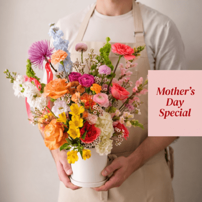 Mother’s Day floral arrangement with coral roses, pink blooms, yellow flowers, soft pastel accents, and seasonal garden flowers arranged in a white ceramic vessel