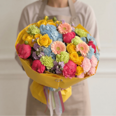 Luxury bouquet with pink peonies, yellow roses, blue hydrangea, peach gerbera daisies, and pastel seasonal flowers wrapped in sage green paper