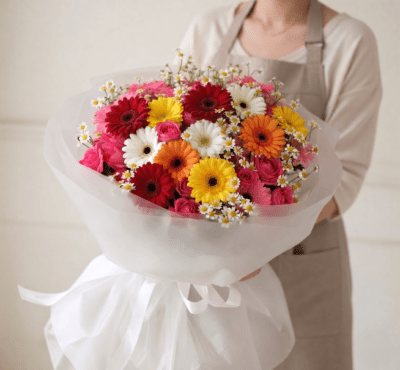 Bright mixed flower bouquet with colourful gerbera-style blooms, pink roses, and delicate white daisy accents wrapped in elegant white paper