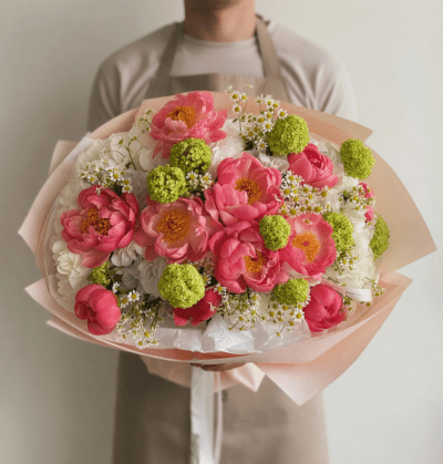 Coral pink bouquet with full blooms, green round accents, and small white flowers wrapped in soft blush paper