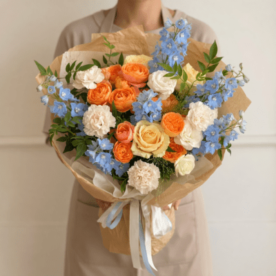 Apricot and cream rose bouquet with white carnations, blue accent flowers, and fresh greenery wrapped in natural kraft paper