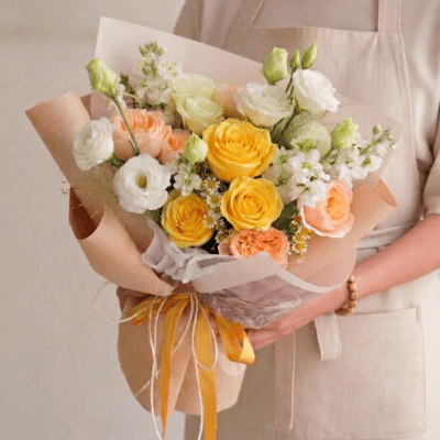 Luxury bouquet with yellow roses, peach roses, white blooms, and delicate daisy accents wrapped in soft neutral paper with ribbon detailing