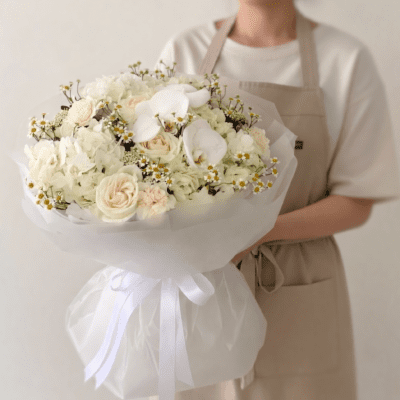 Elegant white bouquet with hydrangea, cream roses, orchid blooms, and delicate chamomile accents wrapped in soft translucent white paper