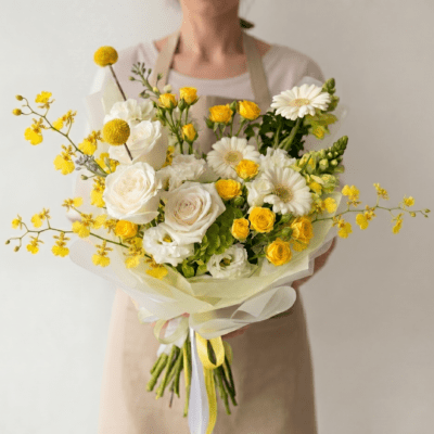 Elegant yellow and white bouquet with ivory roses, yellow spray roses, white gerbera-style flowers, and airy seasonal floral accents wrapped in soft ivory paper