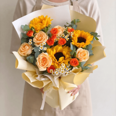Sunflower bouquet with peach roses, orange spray roses, chamomile flowers, eucalyptus greenery, and warm yellow wrapping