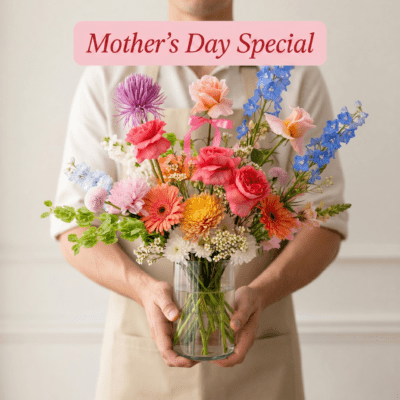 Colourful Mother’s Day vase arrangement with coral roses, pastel flowers, blue delphinium, and fresh greenery in a clear glass vase