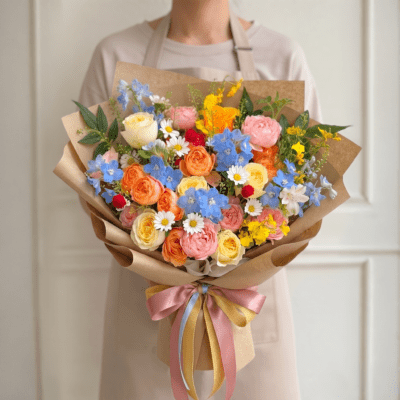 Colourful garden-style bouquet with peach roses, blush blooms, blue flowers, white daisies, and yellow seasonal accents wrapped in kraft paper