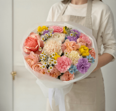 Peach and pastel bouquet with blush roses, cream flowers, chamomile accents, and soft purple blooms wrapped in white paper