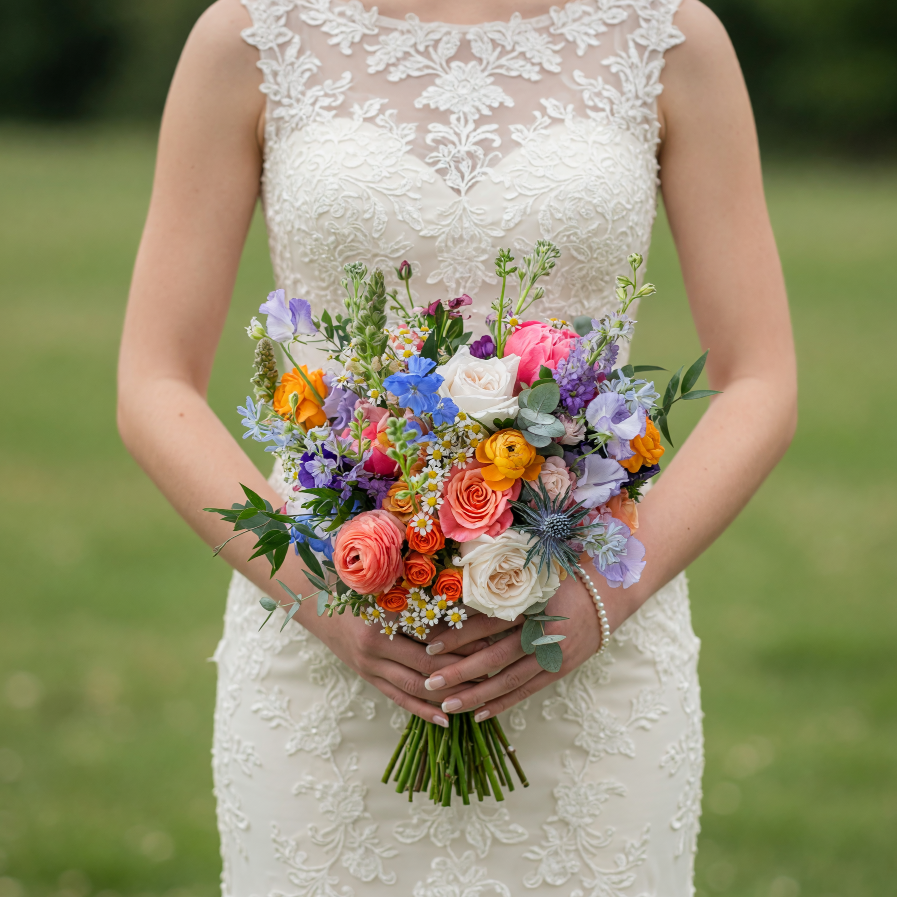 Modern colorful wedding bouquet featuring ivory roses, coral roses, orange ranunculus, blue wildflowers, chamomile, eryngium, eucalyptus, and seasonal greenery.