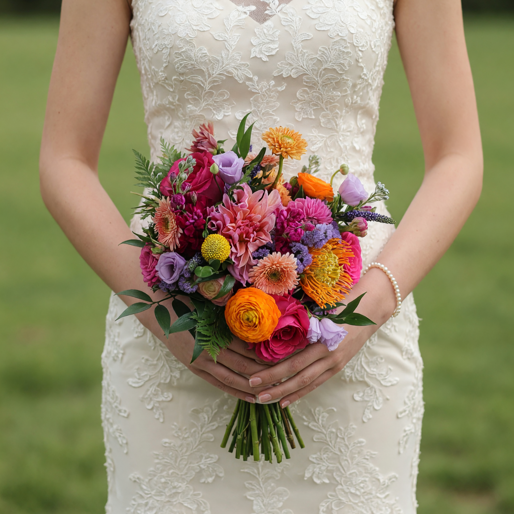 Colorful bouquet for bride with magenta roses, pink dahlias, orange ranunculus, peach gerberas, lavender lisianthus, protea, craspedia, and lush greenery.
