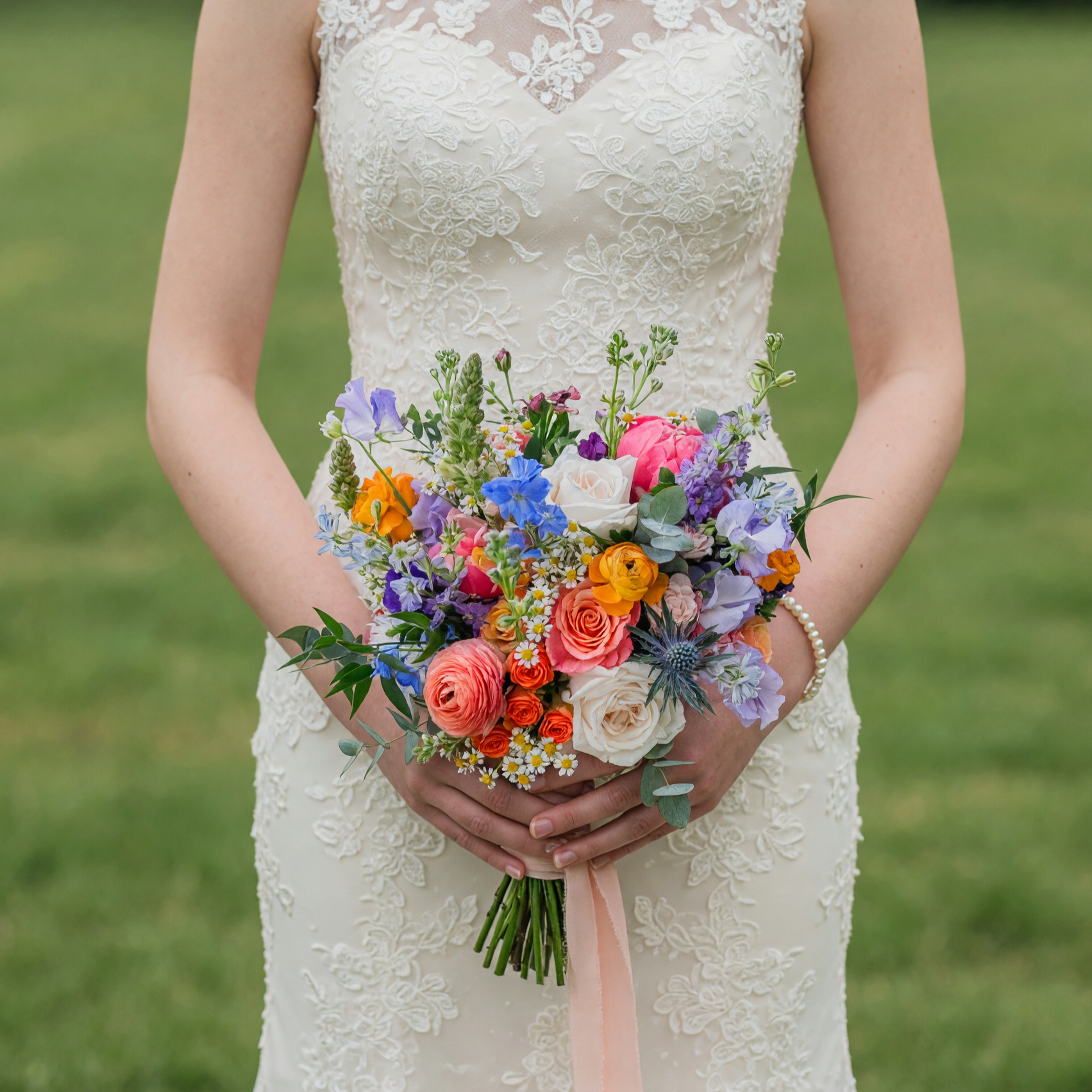 Garden style colorful bouquet featuring peach roses, ivory roses, pink peonies, ranunculus, blue cornflowers, chamomile daisies, eucalyptus, and seasonal wildflowers for a wedding.