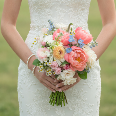 Popular wedding bouquets with coral peonies, peach ranunculus, blush roses, ivory flowers and soft blue blooms held against a white lace bridal gown.
