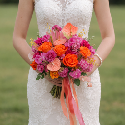 Bridal flowers bouquets with vivid orange roses, pink and magenta flowers, coral anthuriums and flowing peach ribbons held by a bride in a lace gown.