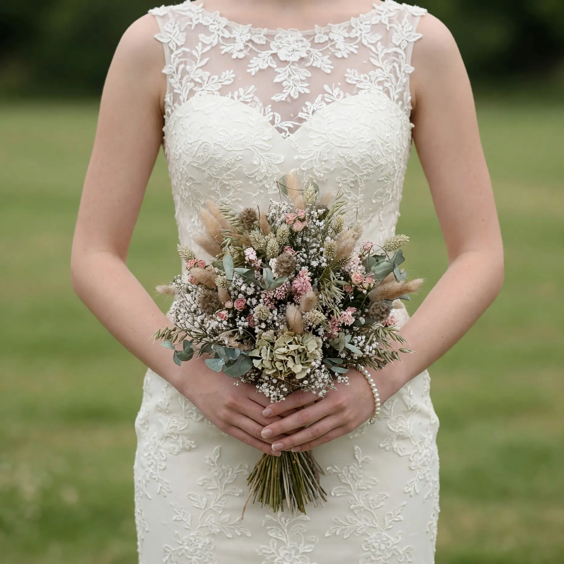 Bride holding a dried flower wedding bouquet with blush pink blooms, tiny ivory blossoms, wheat-like stems, muted greenery, and soft dried grasses.