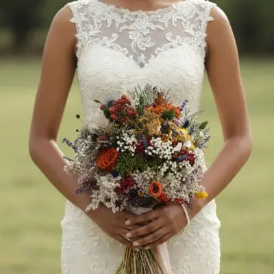 Bride holding a vibrant dried flower wedding bouquet with orange roses, yellow blooms, red berry stems, ivory clusters, blue accents, and greenery.