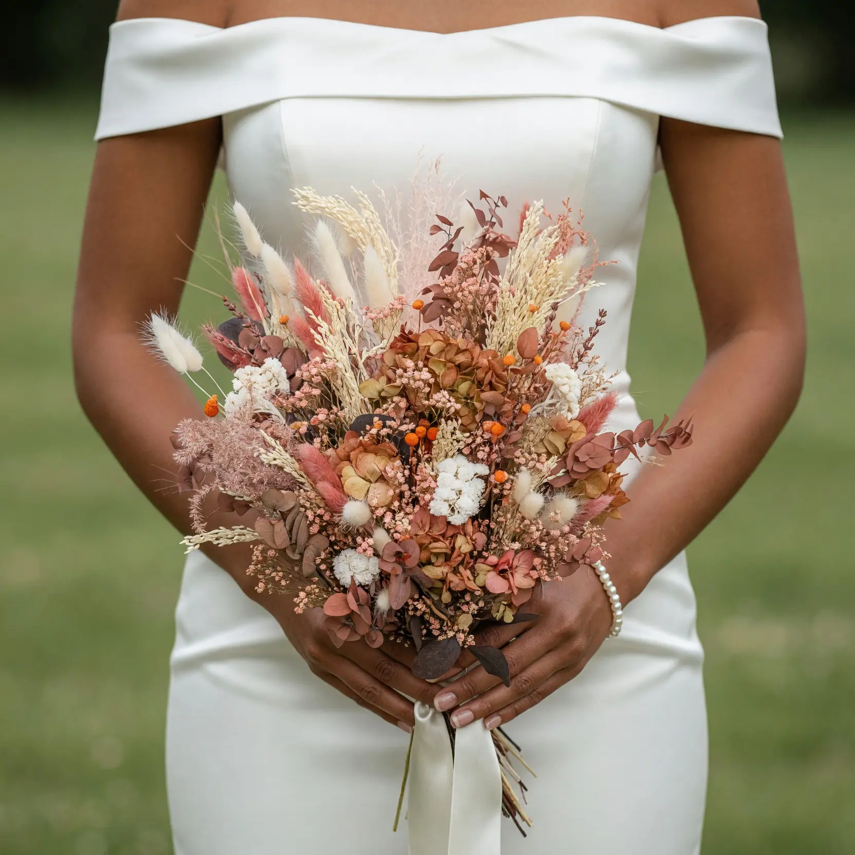 Bride holding a terracotta dried flower wedding bouquet with blush grasses, cream blooms, orange accents, preserved foliage, and ivory ribbon.