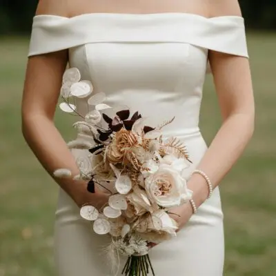 Bride holding a neutral dried flower wedding bouquet with creamy rose-like blooms, translucent seed pods, feathery grasses, beige stems, and burgundy foliage.
