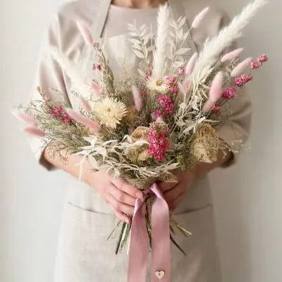 Bride holding a pink dried flower wedding bouquet with cream daisy-like blooms, fluffy pink grasses, pale preserved foliage, and pink ribbon.