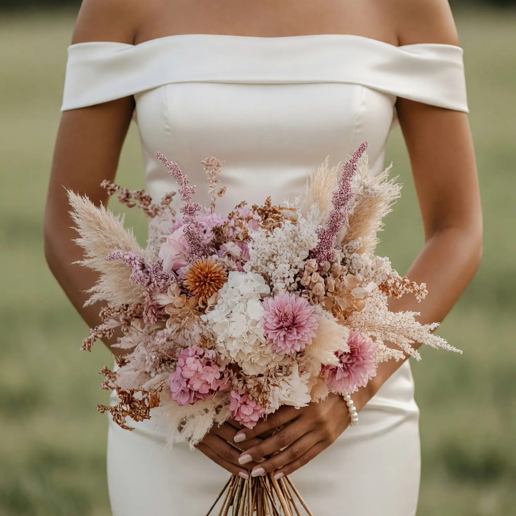 Bride holding a dusty rose dried flower wedding bouquet with pink dahlia-like blooms, cream floral clusters, beige grasses, and mauve stems.