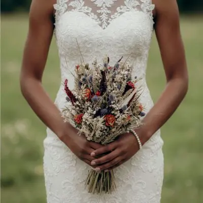 Bride holding a dried flower wedding bouquet with wheat, ivory blossoms, muted blue thistle-like flowers, burgundy foliage, and rust-toned blooms.