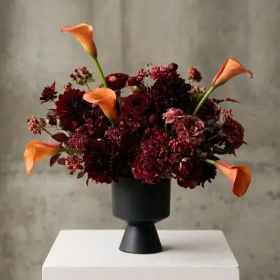 Fall flowers Vancouver arrangement with burgundy blooms, orange calla lilies, berry accents and dark foliage in a modern black pedestal vase.