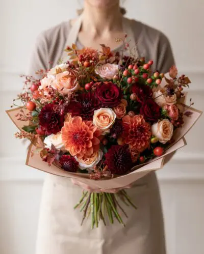 Luxury autumn flowers Vancouver bouquet with burgundy and peach roses, terracotta blooms, red berries and seasonal foliage in neutral wrapping.