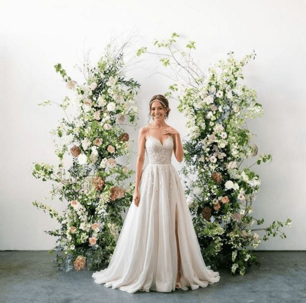 ride in a lace wedding dress standing between tall asymmetrical greenery floral arrangements with white and blush flowers in a minimalist indoor setting.