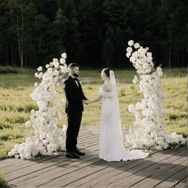 Bride and groom holding hands under a white floral wedding arch with lush blooms during an outdoor ceremony on a wooden platform.