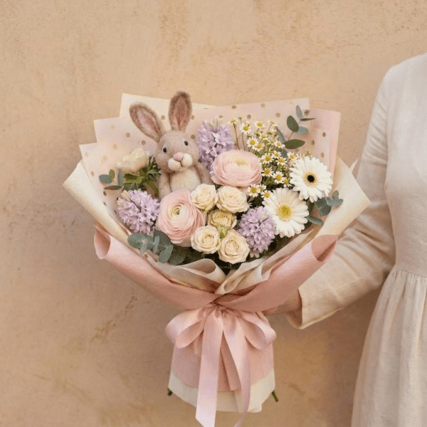 Pastel Easter bouquet with pink and cream flowers, daisies, greenery, and a small bunny wrapped in soft blush paper