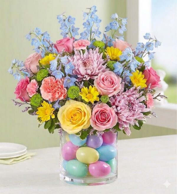 Bright pastel Easter bouquet in a clear glass vase filled with colourful decorative eggs, featuring pink, yellow, blue, and green flowers.