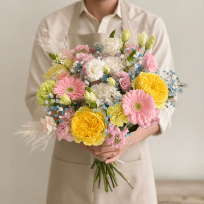Pastel bouquet with yellow roses, pink gerbera, white blooms, and light blue accent flowers in a loose garden style