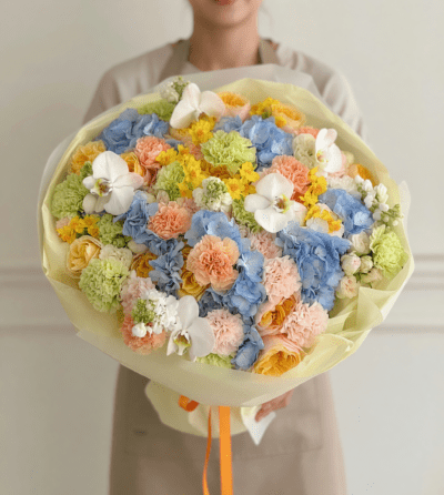Luxury pastel bouquet with blue hydrangea, peach carnations, yellow roses, white orchids, and soft seasonal flowers wrapped in ivory paper