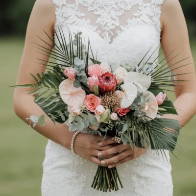 Boho style flowers tropical bridal bouquet with king protea, blush roses, white anthuriums, coral roses, eucalyptus, palm leaves, and tropical greenery