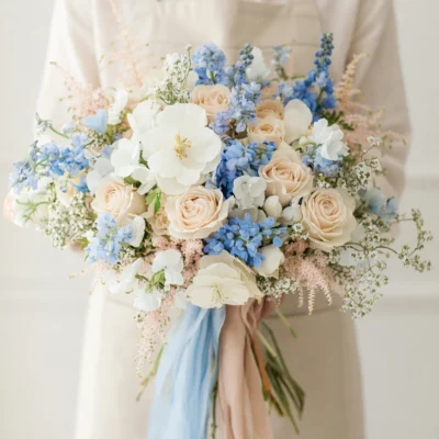 Beach Wedding Bouquets arrangement featuring champagne roses, blue delphiniums, white garden blooms, pale pink accents, delicate filler flowers, and flowing ribbons