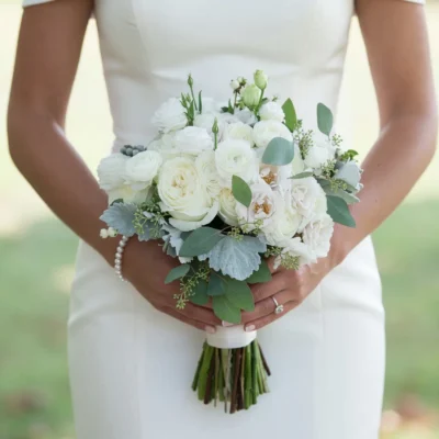 White beach wedding bouquet with ranunculus-like blooms, ivory roses, silver foliage, eucalyptus leaves, and an ivory ribbon wrap.