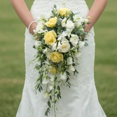 Yellow and white cascading bridal bouquet featuring lemon roses, white blossoms, buds, and fresh greenery against a lace wedding dress.