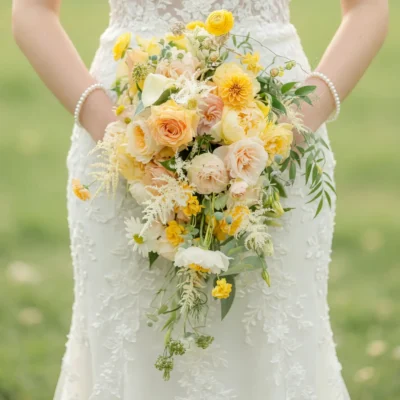 Bride holding a cascading yellow, ivory, and peach wedding bouquet with roses, garden flowers, and trailing greenery.
