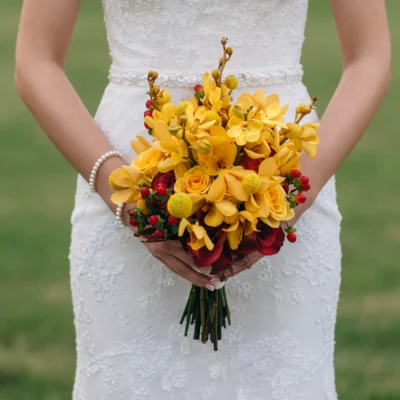 Yellow wedding bouquet featuring golden orchid-like blooms, yellow roses, round button flowers, red berries and burgundy accents held by a bride