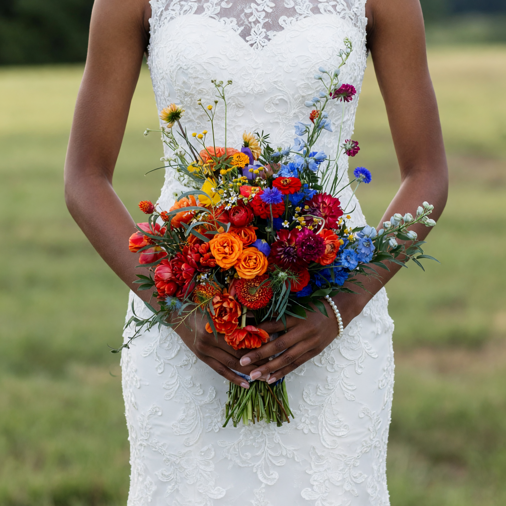 Bridal flowers bouquets with orange roses, vivid blue wildflowers, red blooms, yellow accents and greenery held by a bride in a white lace gown.