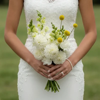 Bridal flowers bouquets with white and ivory dahlias, pale green flower stems, and golden spherical accents held by a bride in an elegant lace wedding dress.