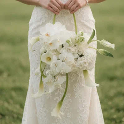 White bridal flowers bouquets design with cascading orchids, calla lilies, textured ivory blooms, and green stems held against a lace wedding dress.