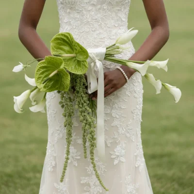 Boho style flowers bridal bouquet with large green tropical blooms, white calla lilies, cascading green texture, and white ribbon against a lace wedding gown.