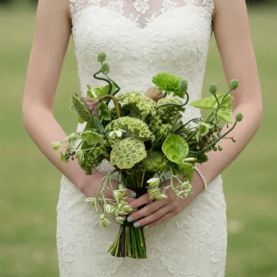 Green bridal bouquet with lotus pods, hydrangeas, botanical flowers, curved stems and lush foliage held by a bride in a lace wedding dress