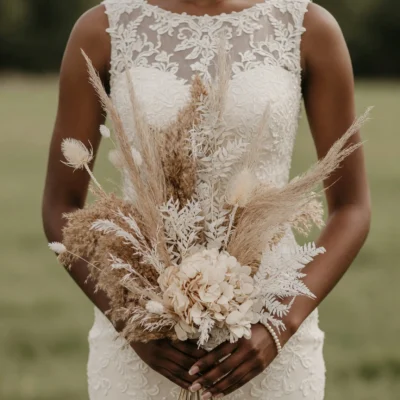 Bride holding an ivory dried flower wedding bouquet with creamy hydrangea-like blooms, pale pampas-like grasses, white fern-like foliage, and warm beige stems.
