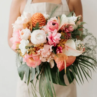 Garden Style Wedding Bouquets Coral Palm combines blush and ivory garden flowers, coral anthurium, warm peach tropical textures, and sweeping palm greenery. Its soft tropical palette is perfect for destination, botanical, coastal, greenhouse, summer, and modern garden weddings.