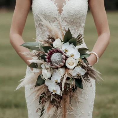 Boho bridal flowers bouquets with white orchids, ivory roses, protea, pampas grass and dark green foliage held by a bride in a lace wedding dress.