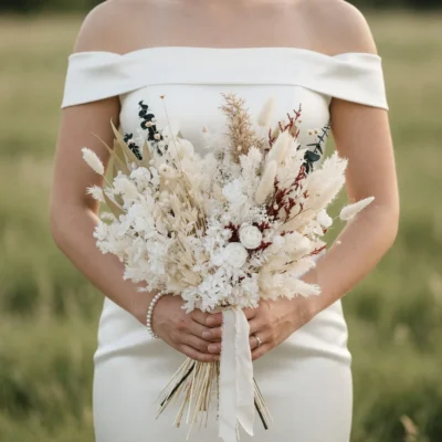 Boho style flowers bridal bouquet with ivory hydrangea blooms, white roses, dried grasses, eucalyptus, and burgundy accents held by a bride.